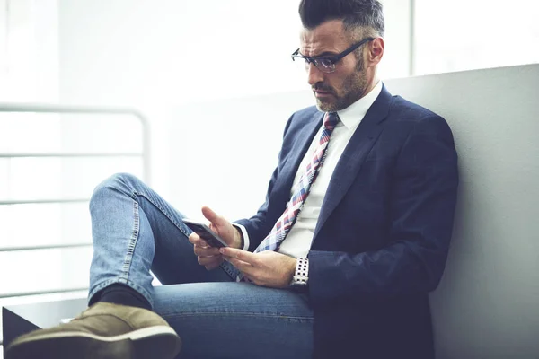 Confident  male director f company dressed in trendy formal outfit and glasses using application on smartphone for banking online, thoughtful businessman reading financial news using wifi connection