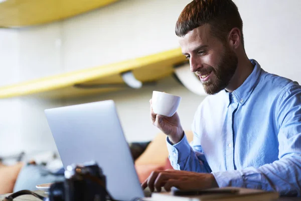 Smiling freelancer copywriter chatting with friends on websites via modern laptop computer device using high speed wireless internet in wifi zone sitting in coffee shop and drinking tasty beverage