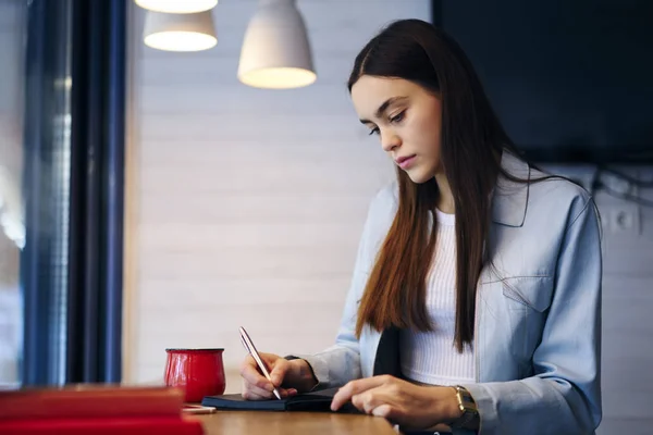 Skilled student dressed in casual outfit writing down notes in notepad and preparing for exams in stylish coworking space.Young woman with brunette hair recording some information in notebook