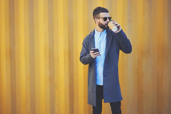 Stylish young male in casual outfit drinking beverage against colorful wall with area for advertising text message,positive bearded man enjoying coffee break in urban setting