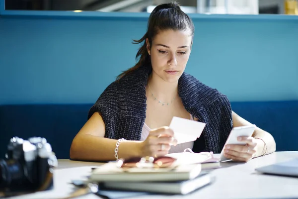 Attractive student holding telephone in hand and reading text information on business card while sitting in coffee shop at table with books.Young woman preparing for training seminar in coworking