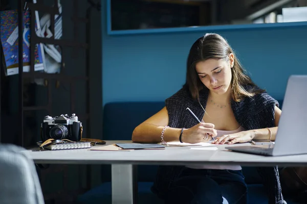 熟练的时髦女孩写下来的文本信息在记事本和做家庭作业坐在桌面上的数字笔记本电脑设备连接到无线互联网 Coworkin 学习的聪明的年轻女子 — 图库照片
