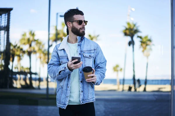 Handsome bearded blogger dressed trendy clothing using application chatting with friends and writing email on cell phone connected to wireless internet,good looking male strolling at free time