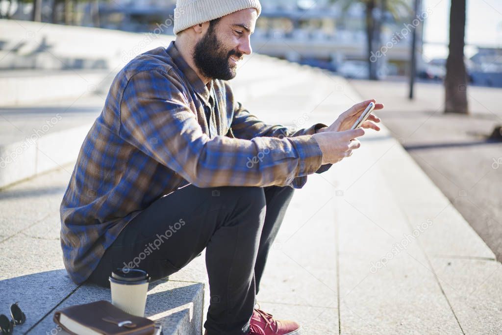 Smiling handsome man in casual outfit blogging in social networks using smartphone and 4G internet connection, bearded hipster guy chatting with friends sitting on city square  during sunny day
