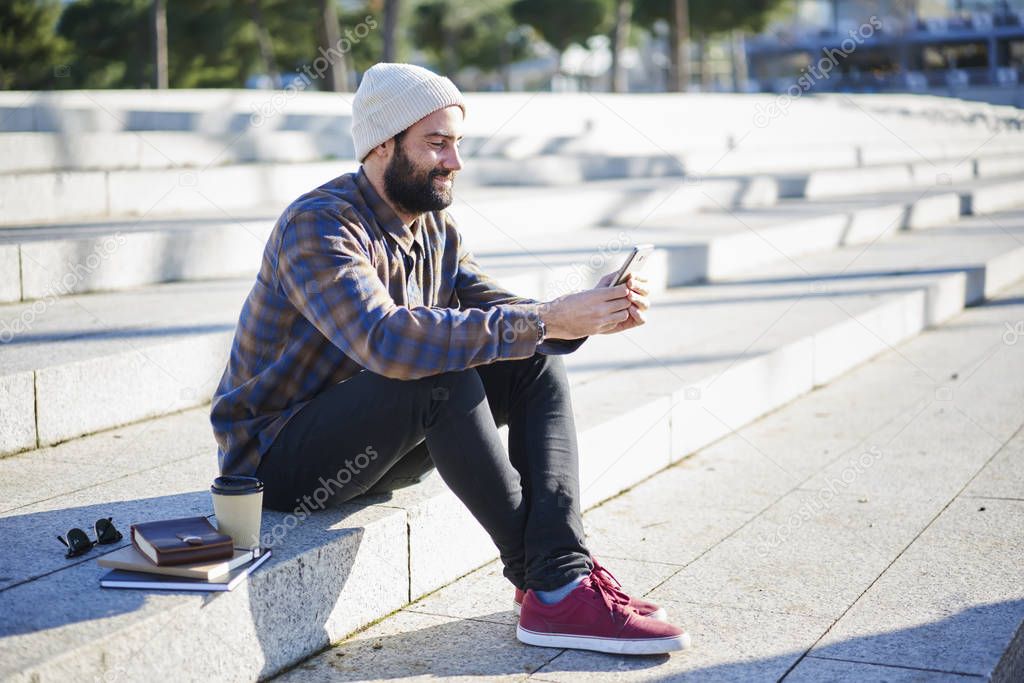 Positive young bearded male student resting outdoors sitting on city square blogging in social networks via phone connected to 4G internet,hipster guy chatting with friend enjoying spring weather