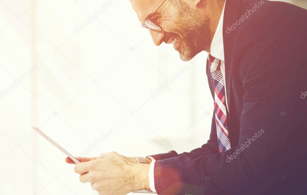 Side view of cheerful male entrepreneur dressed in elegant suit an eyewear checking email on touchpad received feedback with good offer, smiling executive manager satisfied with financial news