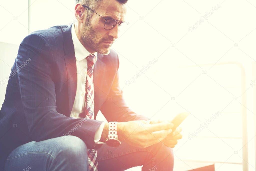 Confident male entrepreneur in trendy eyeglasses dialing number on smartphone for contact banking service, executive manager checking notification on mobile sitting near copy space for advertising