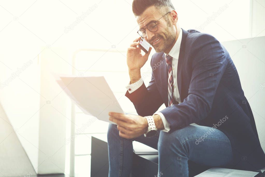 Smiling handsome male entrepreneur satisfied with successful business plan reading report while communicating via smartphone, prosperous mature director talking on phone getting good working offer