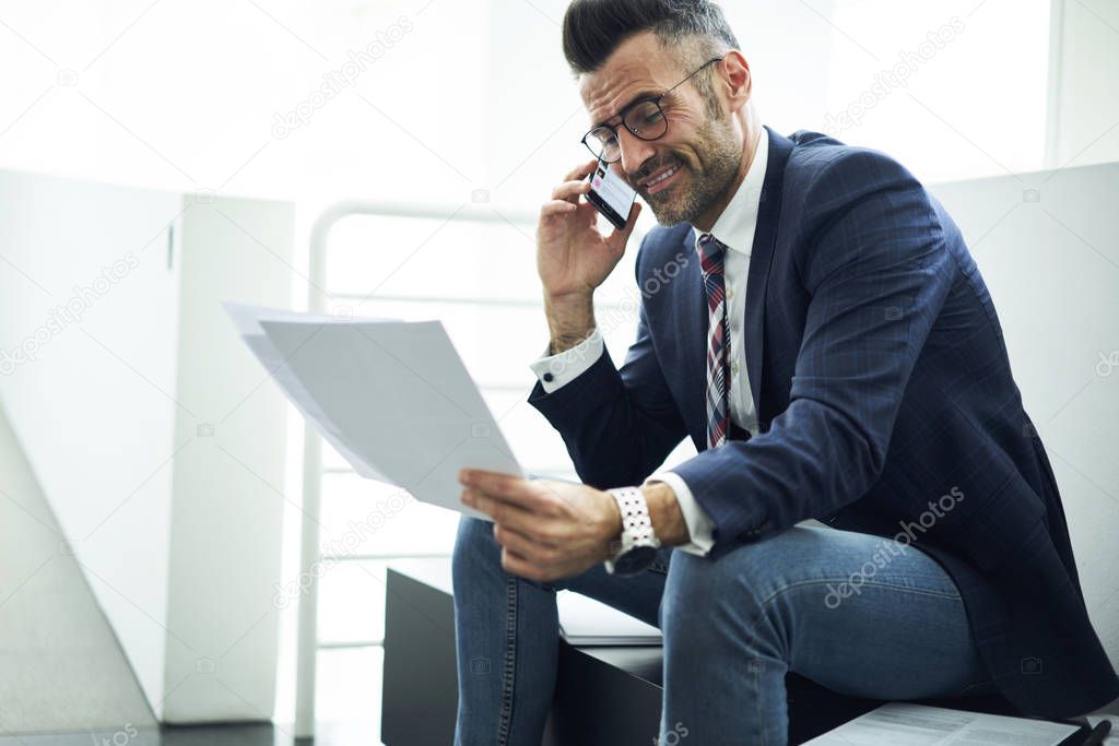Smiling handsome male entrepreneur satisfied with successful business plan reading report while communicating via smartphone, prosperous mature director talking on phone getting good working offer