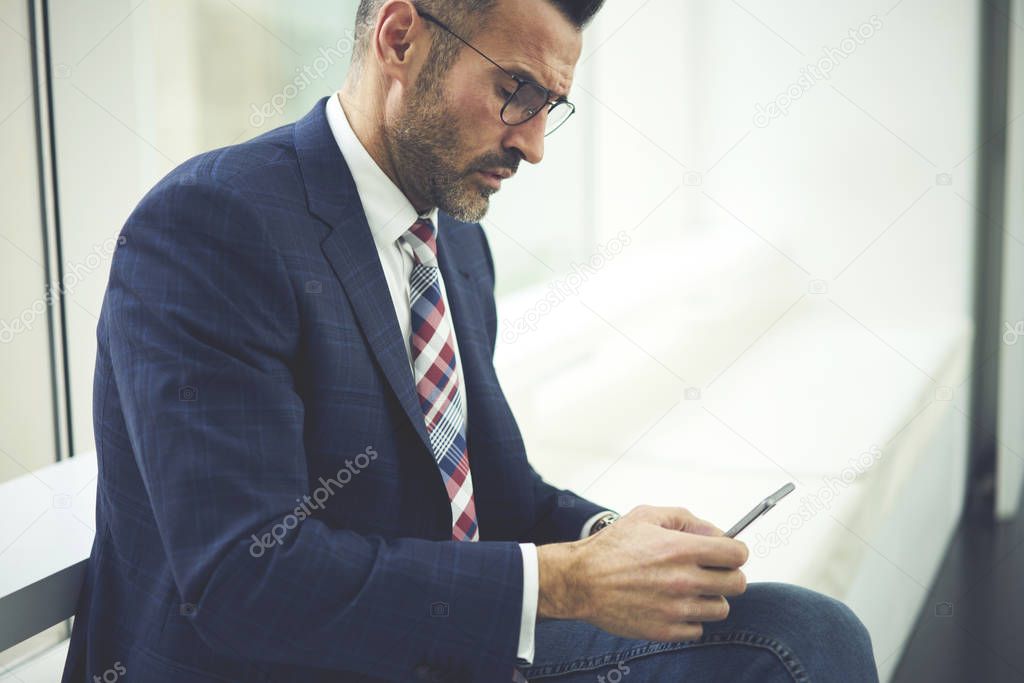Concentrated matured male manager in trendy eyewear checking email box on smartphone sitting near copy space for advertising, confident handsome businessman making payment online via application