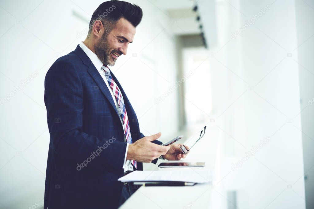 Cheerful mature male executive checking email box on smartphone receiving feedback from business colleagues,positive CEO reading news browsing information via mobile standing on promotional background