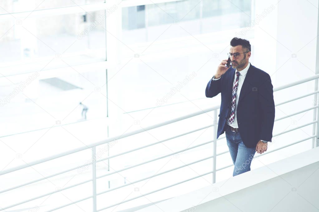 Confident male entrepreneur dressed in formal outfit walking on stairs in white interior office building, experienced matured executive manager talking on mobile phone having business conversation