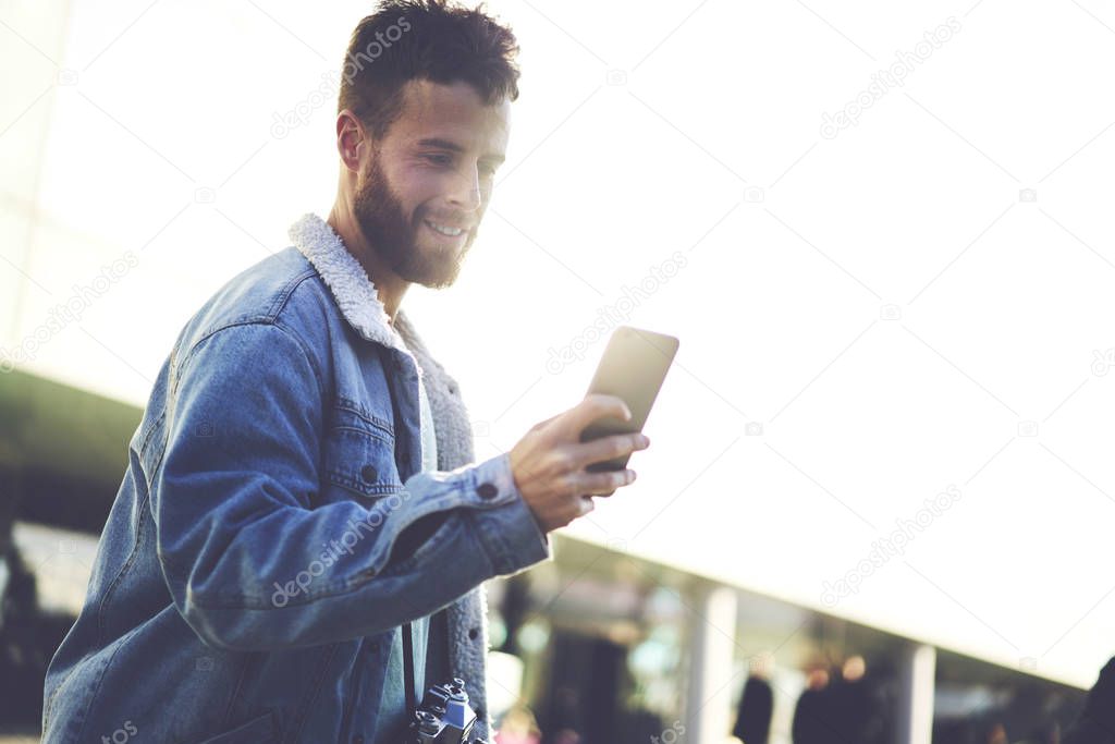 Smiling trendy young student male watching interesting videos in websites for improving skills and knowledge, handsome bearded man in casual outfit writing email on cell phone connected to internet