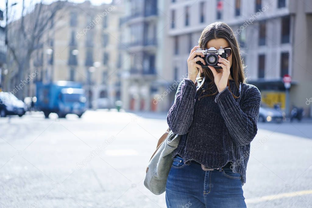 Professional casually dressed female photographer making images of city streets resting during trip.Hipster girl in sunglasses fond of photography using vintage camera strolling on showplaces