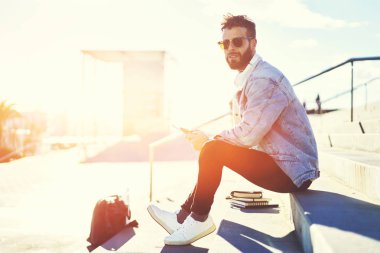 Emin çekici hipster adam dijital tablet sokakta oturan Internet bağlantısı kullanarak üzerinden takipçileri için sosyal ağlar ve mesajlaşma geribildirim arkadaşlarıyla mesajlaşma trendy kıyafetler giymiş