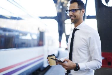Resmi kıyafetler tren sabah iş almak için bekleyen platformda ayakta neşeli genç erkek girişimci işadamı holding taşınabilir pc ve kahve tren istasyonu üzerinde gülümseyen