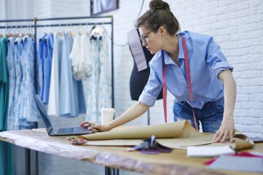 Profesyonel tasarımcı giyim ayakta vasıl okul sırası moda koleksiyonu zemin üzerine dikme için ekipman ile kroki üzerinde çalışırken modern macbook bilgisayar video izlerken gözlük