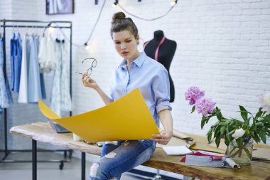 Gözlük elinde tutan ve sarı kroki moda Studio tahta masada oturan bakarak yetenekli tasarımcı. Düşünceli erkek yeni yaz koleksiyonu kendi atelier şık giysi üzerinde çalışıyor 