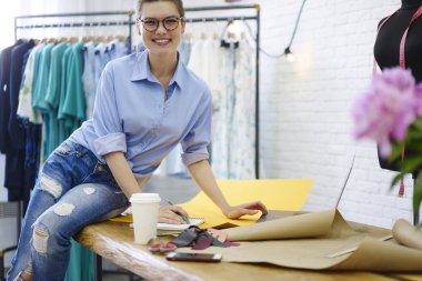 Kameraya bakarak ve giyim mağazası çalışması sırasında gülümseyen süre üstünde okul sırası çizimler ve kahve ile oturan gözlük olumlu modacı. Neşeli tasarımcı oluşturma moda görüntü kendi atelier