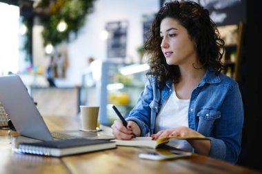 Serin kıvırcık saçlı eğitim webinar izlemek ve genel kavramlar Not Defteri'nde kaydetmek düşünceli yetenekli kız öğrenci. Dizüstü bilgisayarda uygulama kullanırken program planlama Genç Sevimli hippi kız