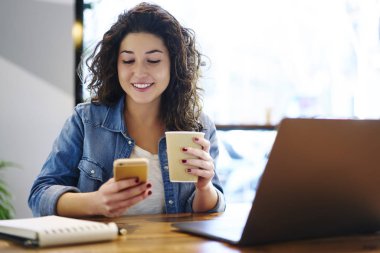 Gülümseyen genç hippi kız aroma kahve tadını çıkarırken bildirim Smartphone cep telefonu ile kontrol modaya uygun kıyafet giymiş. Pozitif sevimli erkek kapalı oturma sırasında cep üzerinde komik fotoğraf görüntüleme