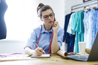 Optik gözlüklü ve modern telefon aracılığıyla müşteriyle iletişim kuran düşünceli profesyonel terzi masaüstünde oturuyor ve kendi giyim mağazasındaki not defterine bilgi yazıyor.