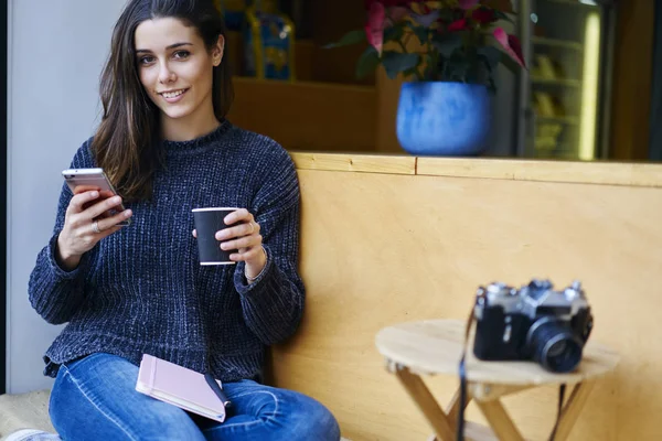 Portrait of charming brunette tourist making booking online via smartphone and internet in roaming.Smiling hipster girl enjoying free time during holiday holding coffee and mobile for texting message