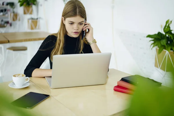 思いやりのある魅力的な女子学生はラップトップに関する情報をブラウズしながら携帯で友達と携帯電話の会話を持っていること 美しい金髪のカフェテリアでの自由時間中に携帯電話で話し — ストック写真