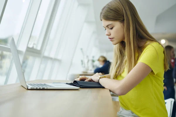 穿着黄色 T恤的沉思的美丽的学生阅读记录写在记事本坐在室内与现代笔记本电脑连接到无线互联网 广告专区 — 图库照片