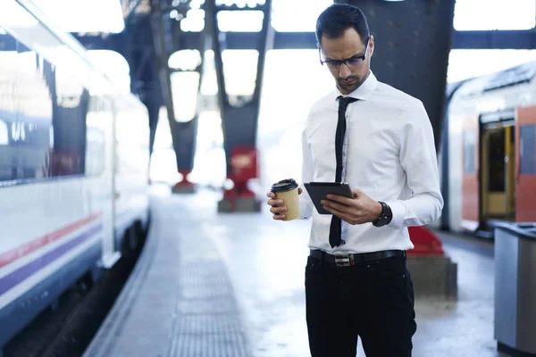 Formally dressed business man in eyewear waiting for train checking transport schedule online on touchpad, concentrated entrepreneur reading news on portable pc holding coffee to go on railway station