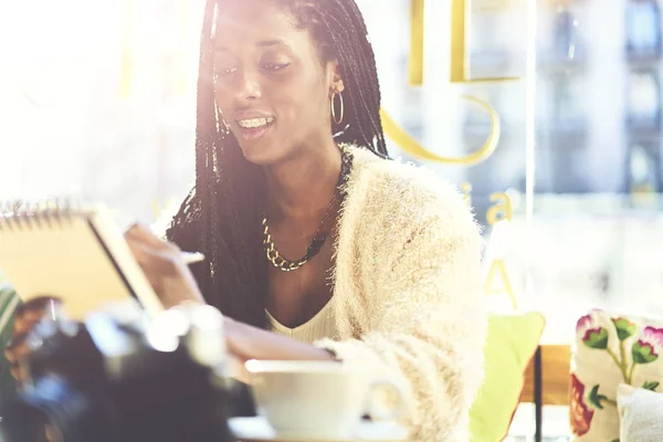 Cheerful afro american graphic designer drawing sketch in notepad enjoying coffee break in cafe interior, positive female journalist with pigtails noting creative ideas for project during leisure