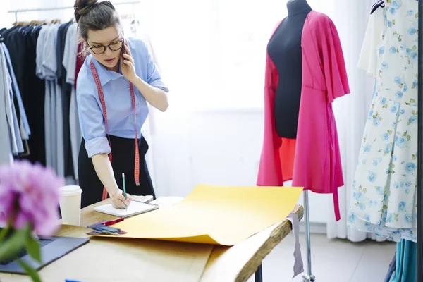 Pensive brunette owner of showroom noting information from phone conversation creating sketch of clothing, concentrated dressmaker writing in notebook measures for tailoring working in studio