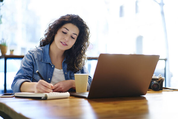 Задумчивая привлекательная женщина-фрилансер с вьющимися волосами, отмечая информацию в блокноте во время работы в лофт-кафе.
