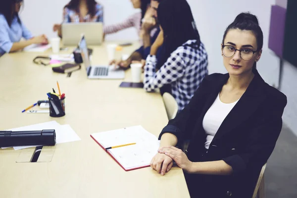 Portrait of charming experienced female employee looking at camera sitting at wooden table while dream team members on background communicating with each other during brainstorming session indoors