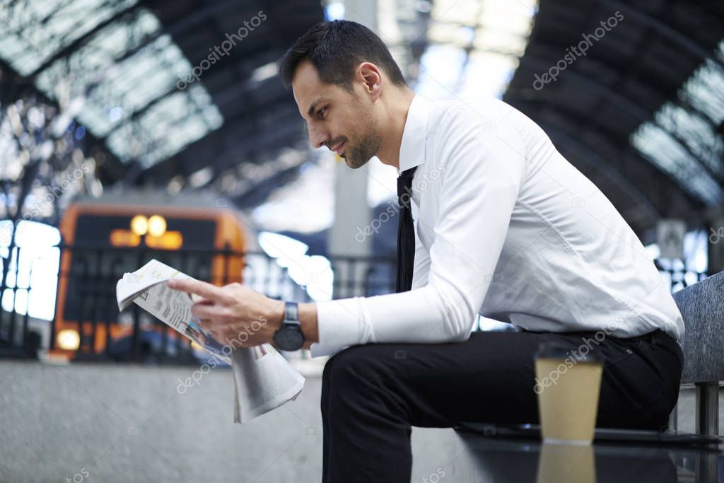 Side view of formally dressed businessman concentrated on reading articles in daily newspaper sitting near platform, prosperous financial manager waiting in railway station for public transport
