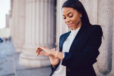 Açık havada şehirde ayakta iken toplantı için zaman yönetimi smartwatch bakarak kurumsal kıyafetleri koyu tenli kadın girişimci. Afro-Amerikan işkadını kol saatleri kontrol