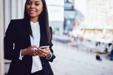 Başarılı bir Afrikalı-Amerikalı kadın girişimci portresi smartphone aygıt ise kameraya gülümseyen ve sokakta ayakta tutan şık resmi takım elbise giymiş. Cep telefonu kullanarak iş kadını