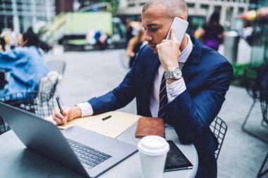Erkek Yöneticisi bilgi yanında açık havada şehir merkezinde oturan smartphone çağrılırken yapım notları şehir kafede, kendine güvenen işadamı oturan telefon görüşme sırasında not defterinde yazılı konsantre