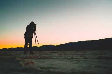 Erkek gazeteci Ölüm Vadisi vahşi topraklarda profesyonel kamera, tripod ve ekipman dus içinde kullanarak Badwater Havzası içinde renkli manzarası çekim adam güzel günbatımı çekim silüeti