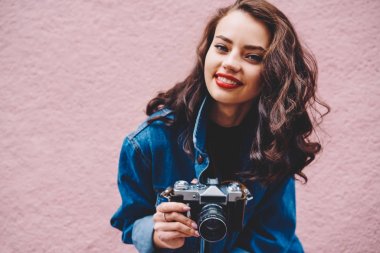 Portre fotoğraf, neşeli esmer kadın Gezgin promosyon pembe vintage fotoğraf makinesi ayakta tutan serbest zaman zevk kıvırcık saç modeli ile hippi kız gülümseyerek, kapatmak