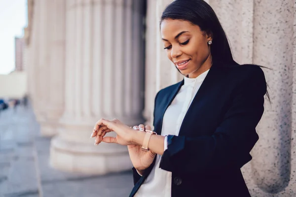 Female entrepreneur with dark skin wearing corporate clothes looking at smartwatch to managing time for meeting while standing outdoors in city. African American businesswoman checking wrist watches