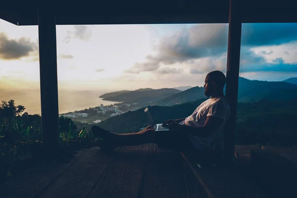 Male blogger writing article on laptop computer while inspiring with beautiful sunset landscape spending vacations with freelance distance work. Thoughtful young man enjoying freedom during travel