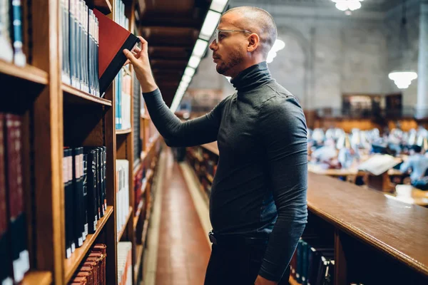 Serious male writer choosing book for reading standing near shelves in old public library, side view young academic student spending time on autodidact selecting literature for increasing knowledge