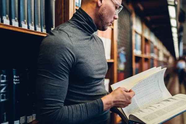 严肃的男性科学家拿着百科全书搜索信息为他的项目站在书架附近 聪明的作家花时间在公共图书馆读经典文学为学习 — 图库照片
