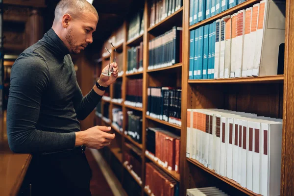 体贴的男性记者摘下眼镜在大学图书馆书架上选择正确的书 沉思的作家为项目花费时间天生搜索信息挑选文学作品 — 图库照片