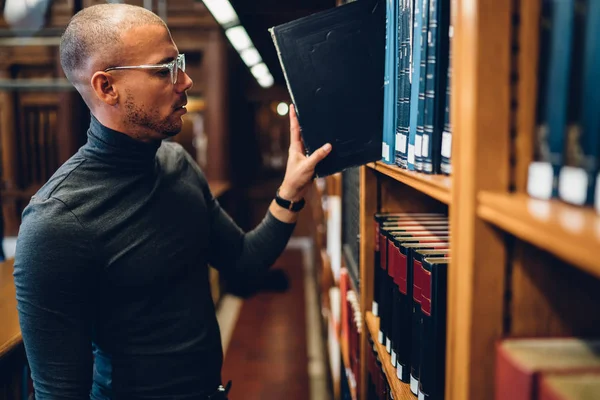 青年男性教授在眼镜选择书为工作与非小说文学 严肃的记者站立在书架子附近图书馆选择百科全书为天生和分析 — 图库照片