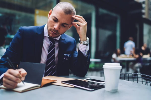 Thoughtful financial manager making notes of budget numbers sitting at table outdoors on urban setting, pensive puzzled businessman writing in notepad analyzing information during coffee break in city