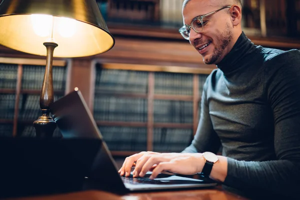 Smiling male writer satisfied with productive work in library typing text of article on laptop for publication on website,cheerful journalist in eyeglasses keyboarding book review for literature page