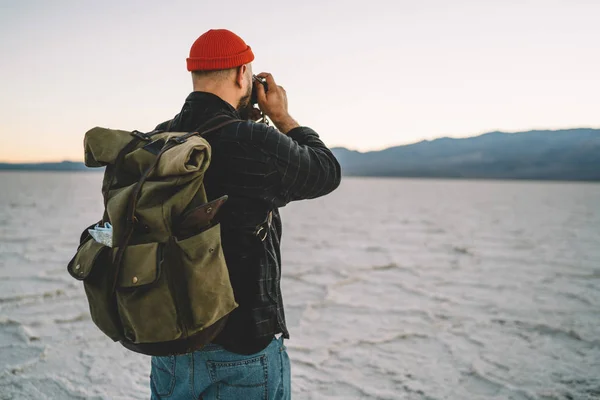 Back view of male photographer traveling and taking picture of unique geological landscape of Death valley,hipster guy with backpack hiking  in Death valley expedition making image of dry surfac