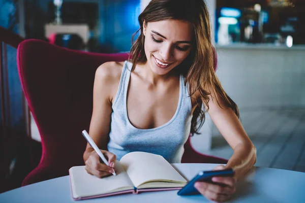 Smiling female noting information from mobile browsing web site sitting in cafe interior, cheerful hipster girl writing in notebook with blank pages waiting for feedback message on smartphone